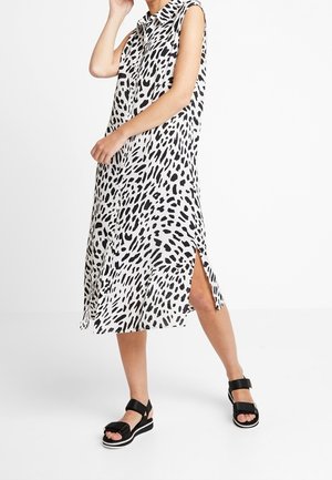 Femme portant une robe midi blanche sans manches avec un motif de points noirs irréguliers et des sandales compensées noires à brides autour de la cheville.