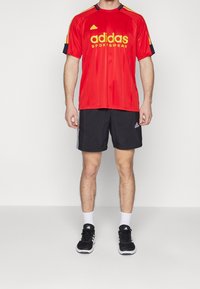 Červený sportovní dres adidas s žlutým logem a akcenty, doplněný černými šortkami s bílými pruhy. Černé sportovní boty a bílé ponožky.