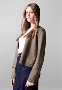 Olivgrüner Cardigan mit gerippten Bündchen und Saum, aus weichem strukturiertem Stoff, getragen über einem hellen Oberteil und kombiniert mit dunklen Hosen.