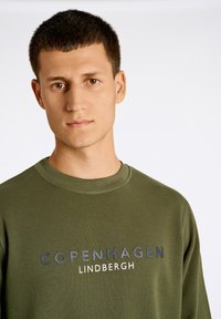 Olivgrön sweatshirt med rund hals, försedd med en tryckt "COPENHAGEN LINDBERGH"-logotyp i kontrasterande färger på framsidan. Slät textur.