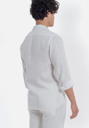 Mężczyzna z kręconymi włosami, ubrany w białą lnianą koszulę z długim rękawem i dopasowane białe spodnie, stojący na jednolitym jasnym tle.