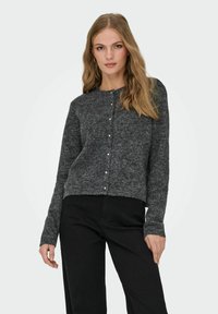 Ung kvinna med långt blont hår, iklädd en mörkgrå knappad cardigan och svarta byxor, stående mot en enkel vit bakgrund.