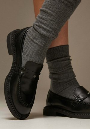 Black leather loafers with decorative stitching, paired with ribbed gray socks, set against a neutral background.