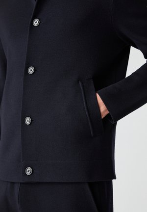 Chaqueta de punto navy con cuello, cinco botones y un bolsillo lateral. La tela tiene un acabado texturizado con puños y dobladillo acanalados.
