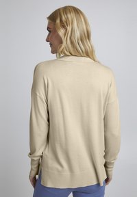 Beige hosszú ujjú pulóver laza szabással, puha tapintással, oldalsó hasítékokkal az alján és klasszikus gallérral, világoskék nadrág felett viselve.