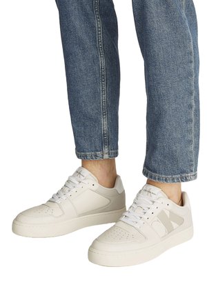 Sneakers bianche basse con punte traforate e logo "CK", indossate con jeans blu risvoltati da una persona in piedi.