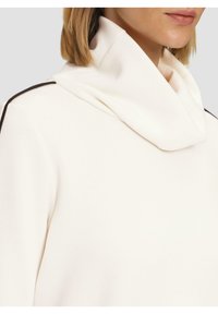 Svetr s kapucí, off-white barvy, vyrobený z jemného materiálu. Vyznačuje se tmavě hnědými akcenty na ramenou pro kontrast.