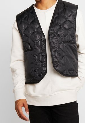 Homme portant un gilet matelassé noir à boutons par-dessus un sweat-shirt blanc à manches longues et un pantalon noir, visible de la poitrine à la taille.