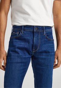 Blue denim jeans with a regular fit, featuring contrast stitching, five-pocket design, and a metal button closure. Worn with a white shirt.