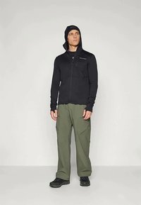 Άνδρας που φοράει μαύρο φούτερ με φερμουάρ και κουκούλα πάνω από μαύρο καπέλο, ελαιοπράσινα cargo παντελόνια και μαύρα παπούτσια, στέκεται έναντι απλής φόντου.