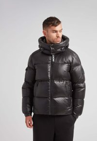 Giacca a vento nera con colletto alto, tasche con zip e texture lucida. Presenta cordini regolabili e un logo sul davanti.