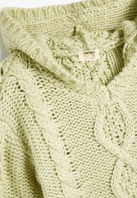 Stickad huvtröja i ljusgrön, med en grov textur, kabelmönster och en vikkrage. Tagg synlig med storleksdetaljer.