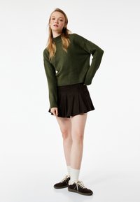 Zielono-oliwkowy sweter dzianinowy z wysokim kołnierzem, w połączeniu z czarną plisowaną mini spódnicą. Modelka nosi ciemne trampki i białe skarpetki.