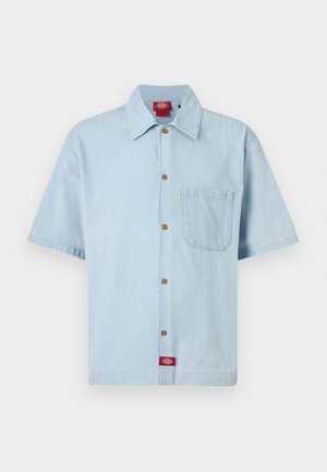 Chemise bleu clair à manches courtes avec poche poitrine, col classique, boutons marron, et étiquette de marque rouge sur l'ourlet.