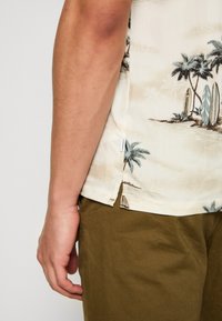 Bras masculin reposant à côté d'un pantalon vert olive, portant une chemise crème avec un imprimé de palmier et de planche de surf près de l'ourlet.
