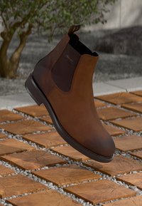 Botín Chelsea de cuero marrón con paneles laterales elásticos, textura suave y puntera redondeada. Presenta un tacón bajo y un sutil logo en el lateral.