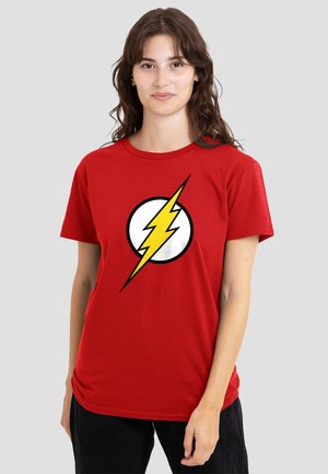 Young woman with long brown hair wearing a red t-shirt featuring a yellow lightning bolt inside a white circle, standing against a gray background.