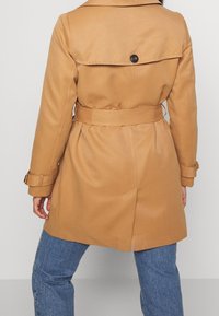 Béžový trench coat s páskem, zadním rozventilovaným dílem a knoflíkovým detailem na límci. Má dlouhé rukávy a je kombinován s modrými denimovými džíny.