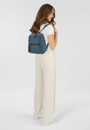 Mochila azul con tela texturizada y acentos lisos, presenta correas ajustables y un bolsillo frontal con cremallera. El modelo lleva pantalones claros.
