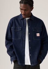 Veste en denim bleu foncé avec deux poches poitrine, coutures contrastantes et fermeture à boutons. Portée sur un t-shirt blanc.