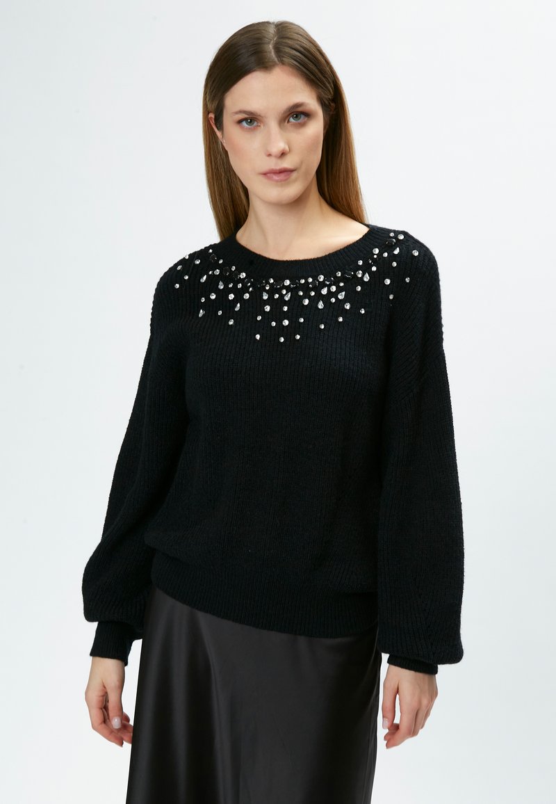 Schwarzer Strickpullover mit rundem Ausschnitt, verziert mit klaren Edelsteinen, die in einem dekorativen Muster auf der Vorderseite angeordnet sind.