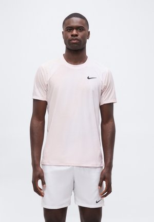Mann som står iført lys rosa Nike T-skjorte og hvite Nike shorts mot ensfarget lys grå bakgrunn.