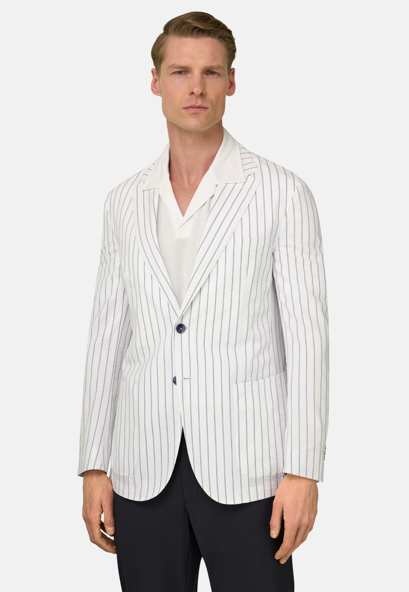 Homme portant un blazer blanc à simple boutonnage avec de fines rayures verticales, une chemise blanche et un pantalon noir, debout devant un fond uni.