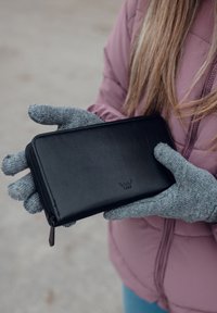 Black leather wallet with a zip closure, featuring a logo, held by a person wearing grey knit gloves, against a neutral background.