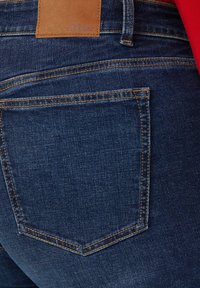 Ciemnoniebieskie dżinsy z widoczną tylną kieszenią z jasnopomarańczowym przeszyciem. Skórzana metka z logo umieszczona na pasku od spodni.