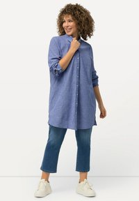 Camicia a righe blu e bianche con colletto, maniche lunghe e polsini arrotolati, abbinata a jeans blu a vita alta e sneakers bianche.