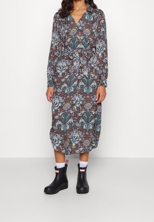 Frau trägt ein langärmliges, geblümtes Kleid mit Gürtel, kombiniert mit schwarzen Regenstiefeletten und weißen Socken, steht vor einem einfarbigen Hintergrund.