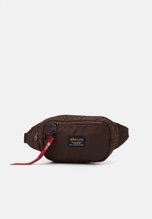 Brązowa materiałowa torba na pas z przednią kieszenią na suwak, czarną naszywką z logo Alpha Industries i czerwoną materiałową zawieszką zamka.