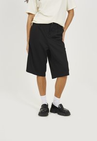 Black bermuda shorts made of smooth fabric, featuring a relaxed fit and side pockets, styled with a cream T-shirt and chunky black loafers.