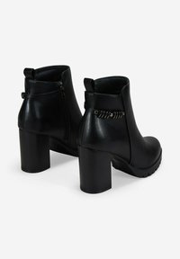 Botas de piel negra hasta el tobillo con tacones altos y gruesos, cremalleras laterales y correas decorativas con acentos metálicos alrededor de la parte posterior.