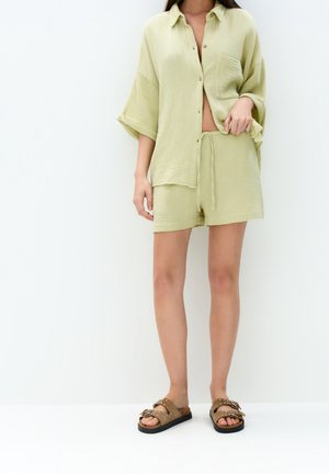 Kvinde iført løs, lysegrøn, tekstureret skjorte med knapper og matchende shorts med brune sandaler med nitter mod hvid baggrund.