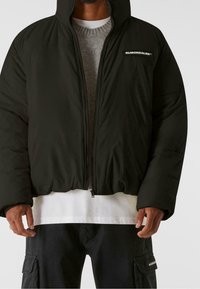 Giubbotto piumino nero con cappuccio, dotato di chiusura lampo e patch con logo sul petto. Materiale spesso con una texture liscia.