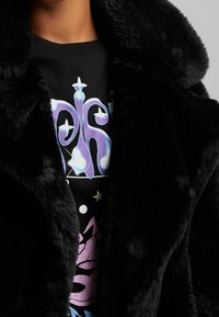 Person wearing a black faux fur coat over a black shirt with abstract purple, blue, and green graphic designs and star shapes.