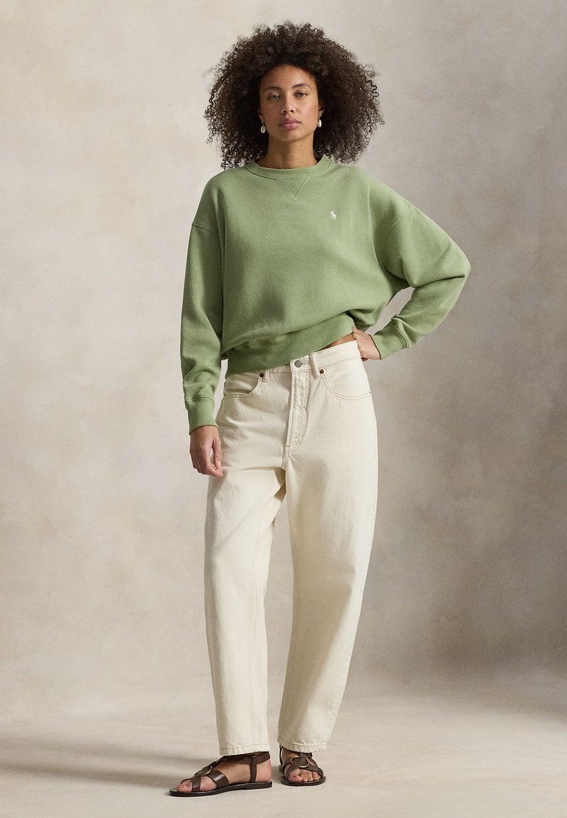 Green oversized sweatshirt with ribbed cuffs, light beige loose-fitting jeans, and brown strappy sandals, on a neutral background.