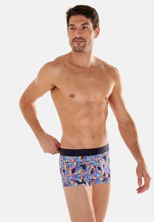 Mężczyzna bez koszulki stoi, nosi kolorowe bokserki w kwiatowy wzór, jedną ręką reguluje talię, patrząc na bok na jednolitym tle.