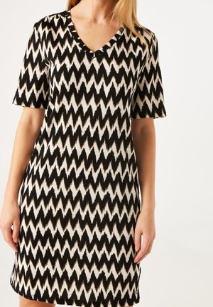 Woman wearing a black and white zigzag patterned short-sleeve dress with a V-neck, standing against a plain white background.