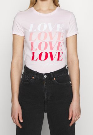 Kobieta w jasnoróżowej koszulce z napisem "LOVE" powtarzanym w odcieniach czerwieni oraz frazą "is all you need", zestawiona z czarnymi dżinsami.