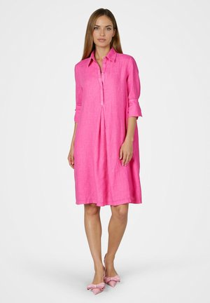 Woman wearing a bright pink knee-length shirt dress with three-quarter sleeves and matching pink pointed shoes with bows.