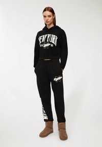 Czarna, krótka bluza z kapturem z nadrukiem "NEW YORK 1991", zestawiona z czarnymi spodniami dresowymi z białym napisem "NEW YORK" oraz brązowymi zamszowymi botkami do kostek.