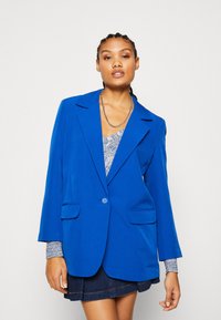 Modra blazer s strukturiranim dizajnom, z enim gumbom, dvema sprednjima žepoma in ostrimi reverji. Nosi se preko vzorčaste bluze in kavbojskih kril.