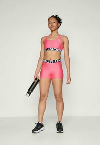 Rózsaszín sportmelltartó és rövidnadrág szett széles fekete derékpánttal, rajta az "UNDER ARMOUR" felirattal. Fekete vizespalackot tart. Fekete sportcipő.