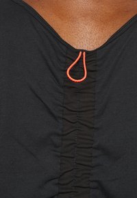Gros plan sur un haut noir froncé avec un décolleté en V et un détail boucle orange vif au centre du décolleté.