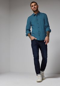 Tyrkysová košile s dlouhým rukávem a ohrnutými rukávy, tmavé denimové džíny a bílé tenisky. Jednoduchý design bez viditelných vzorů nebo log.