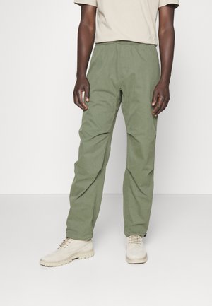 Homme portant des chaussures beige clair, un pantalon vert olive à taille élastique et une chemise beige à manches courtes, debout devant un fond uni.