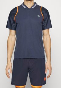 Marinblå sportspolo med meshärmar, orange detaljer och en krage. Matchas med marinblå shorts med orange kant och logotypdetalj.