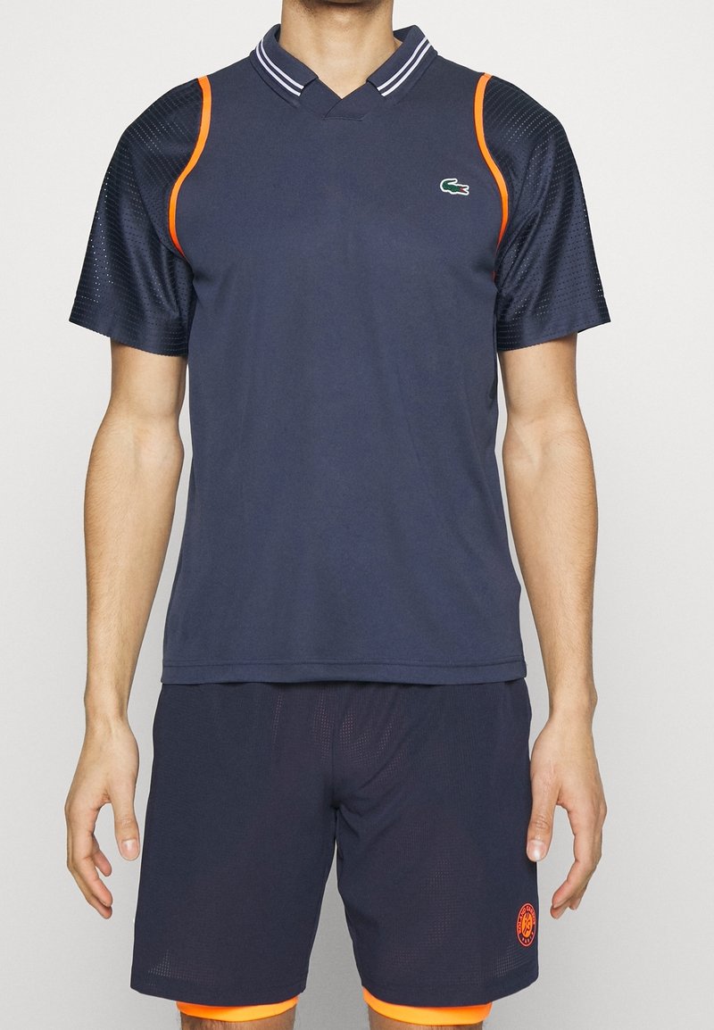 Marinblå sportspolo med meshärmar, orange detaljer och en krage. Matchas med marinblå shorts med orange kant och logotypdetalj.